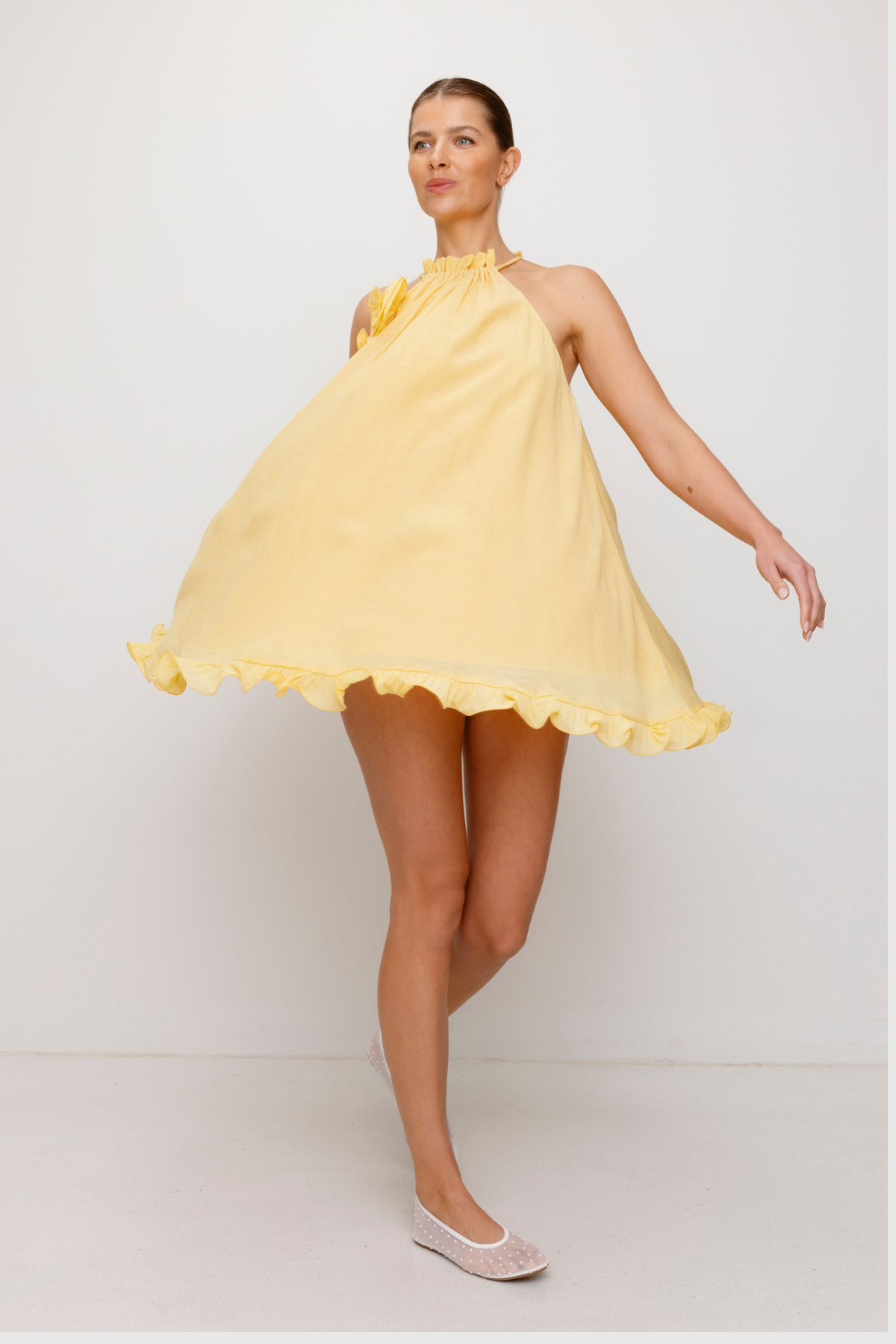 Flowy trapeze mini dress in butter, yellow, featuring rosette neckline detail, tie neck, frilled hem, and easy pull-on fit.