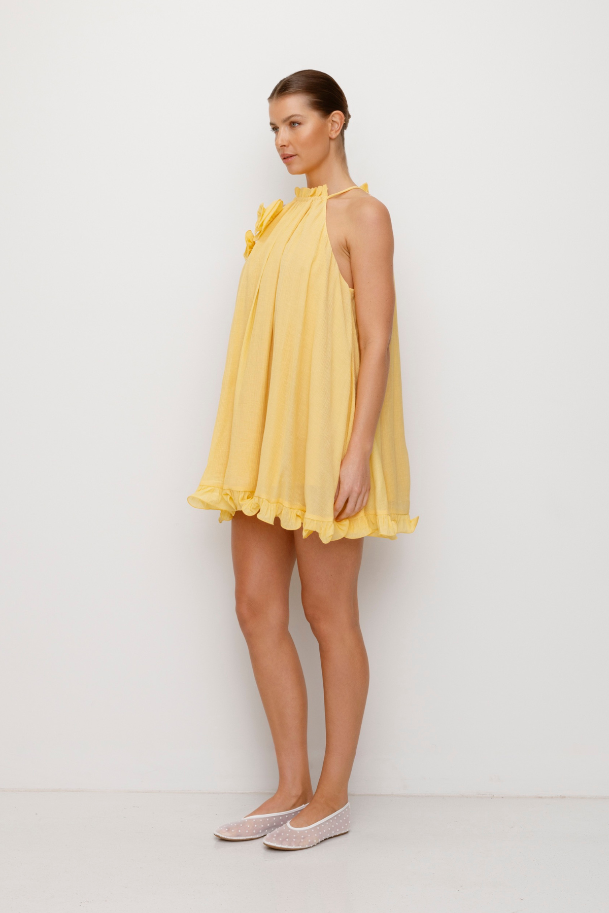 Flowy trapeze mini dress in butter, yellow, featuring rosette neckline detail, tie neck, frilled hem, and easy pull-on fit.