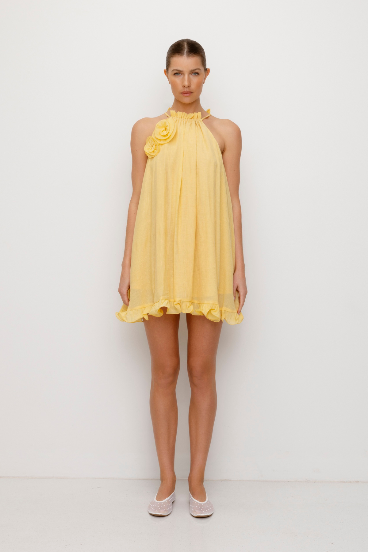 Flowy trapeze mini dress in butter, yellow, featuring rosette neckline detail, tie neck, frilled hem, and easy pull-on fit.