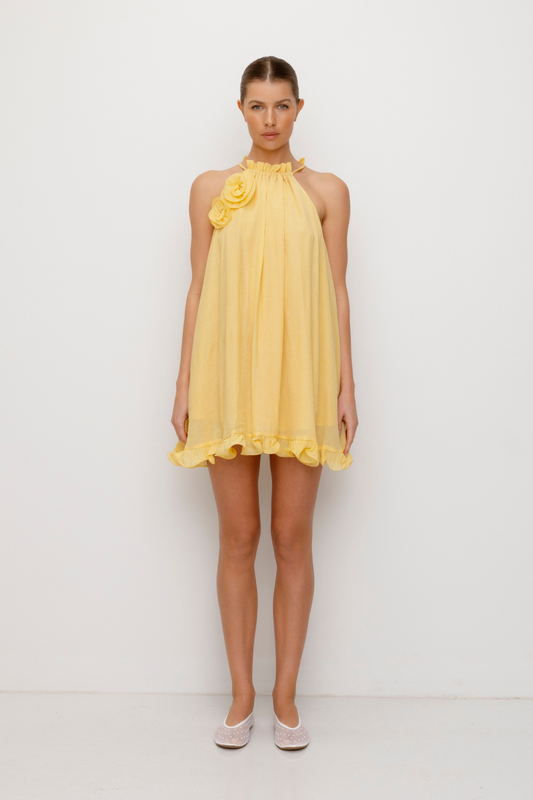 Flowy trapeze mini dress in butter, yellow, featuring rosette neckline detail, tie neck, frilled hem, and easy pull-on fit.