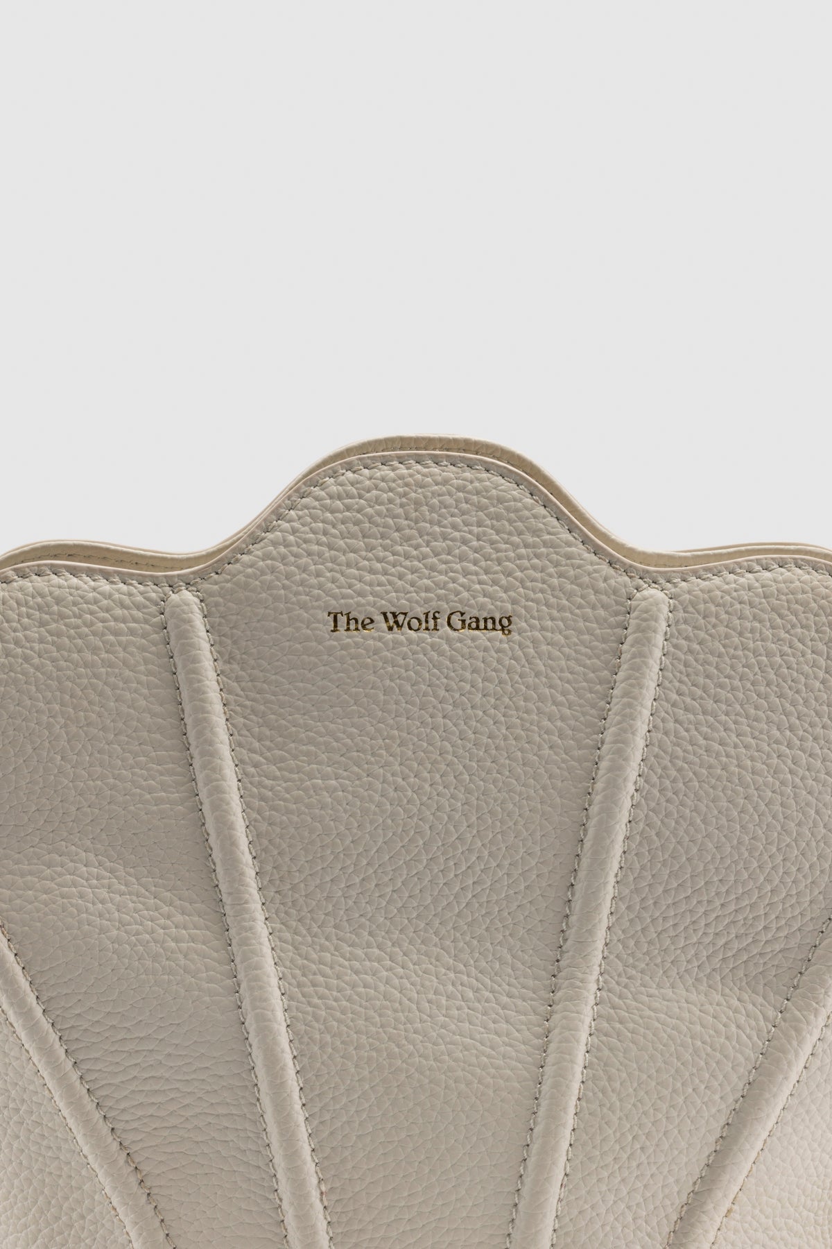 Luxury pebbled leather Suri Bag shell-shaped clutch in Ivory, with scalloped edge, gold zip, detachable crossbody strap, and ridged detail.