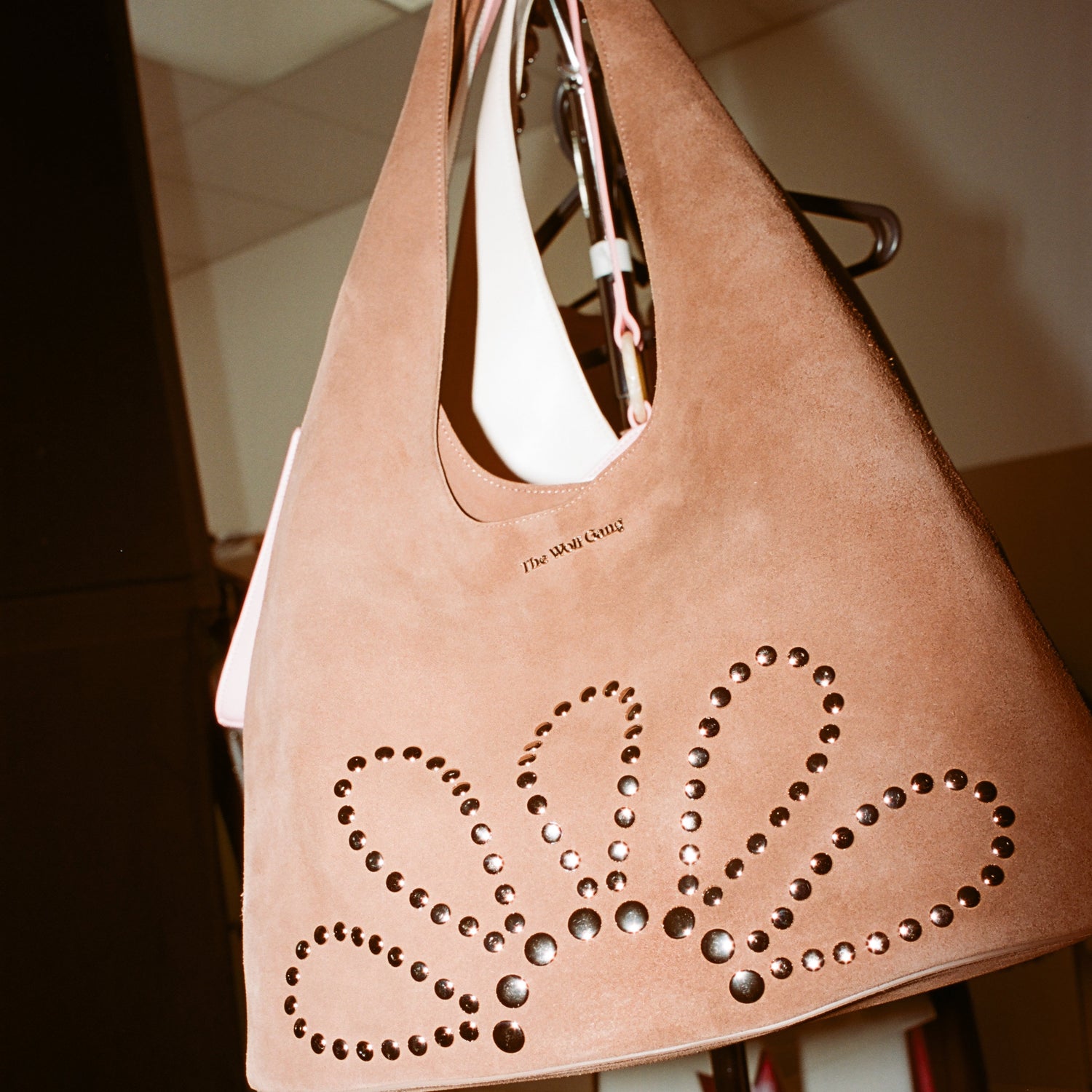 A large tan suede shoulder bag with studded floral cutout detailing, hanging on a display stand in an indoor showroom.
