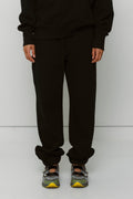 Model displays black sweatpants and matching sweatshirt with Asics sneakers against a white wall.
