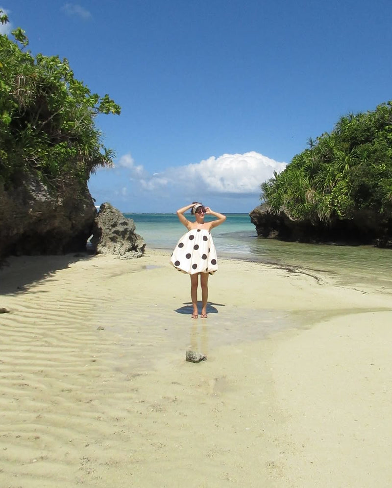 A woman stands on a sandy beach between rocky cliffs and green vegetation, wearing a white dress with black polka dots, facing the ocean under a bright blue sky.