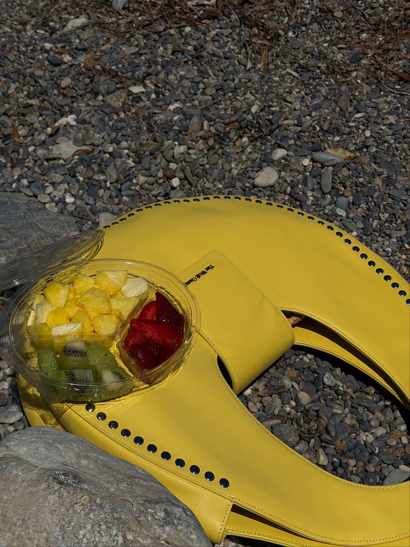A bright yellow designer handbag with a clear dome container holding sliced fruit, placed on a bed of gray gravel stones.