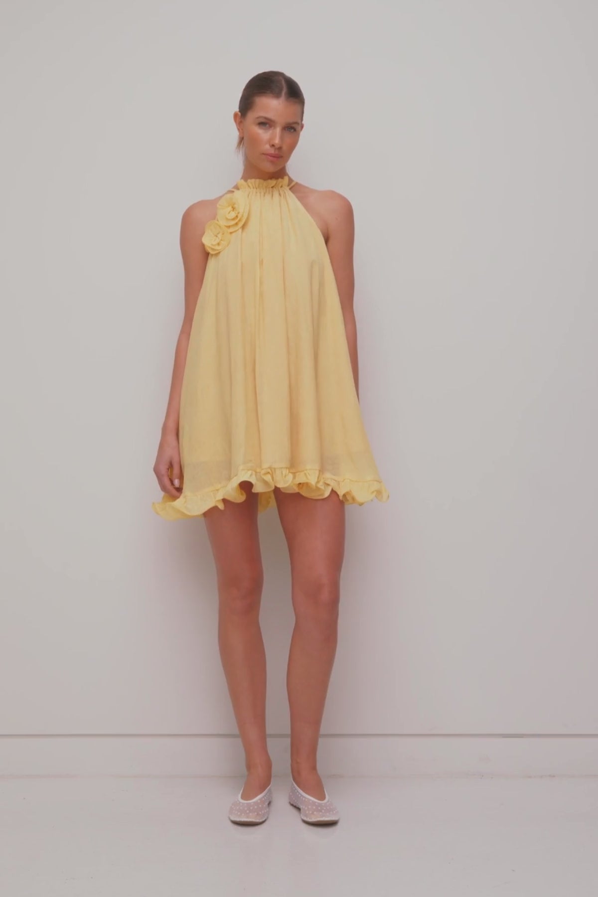 Flowy trapeze mini dress in butter, yellow, featuring rosette neckline detail, tie neck, frilled hem, and easy pull-on fit.
